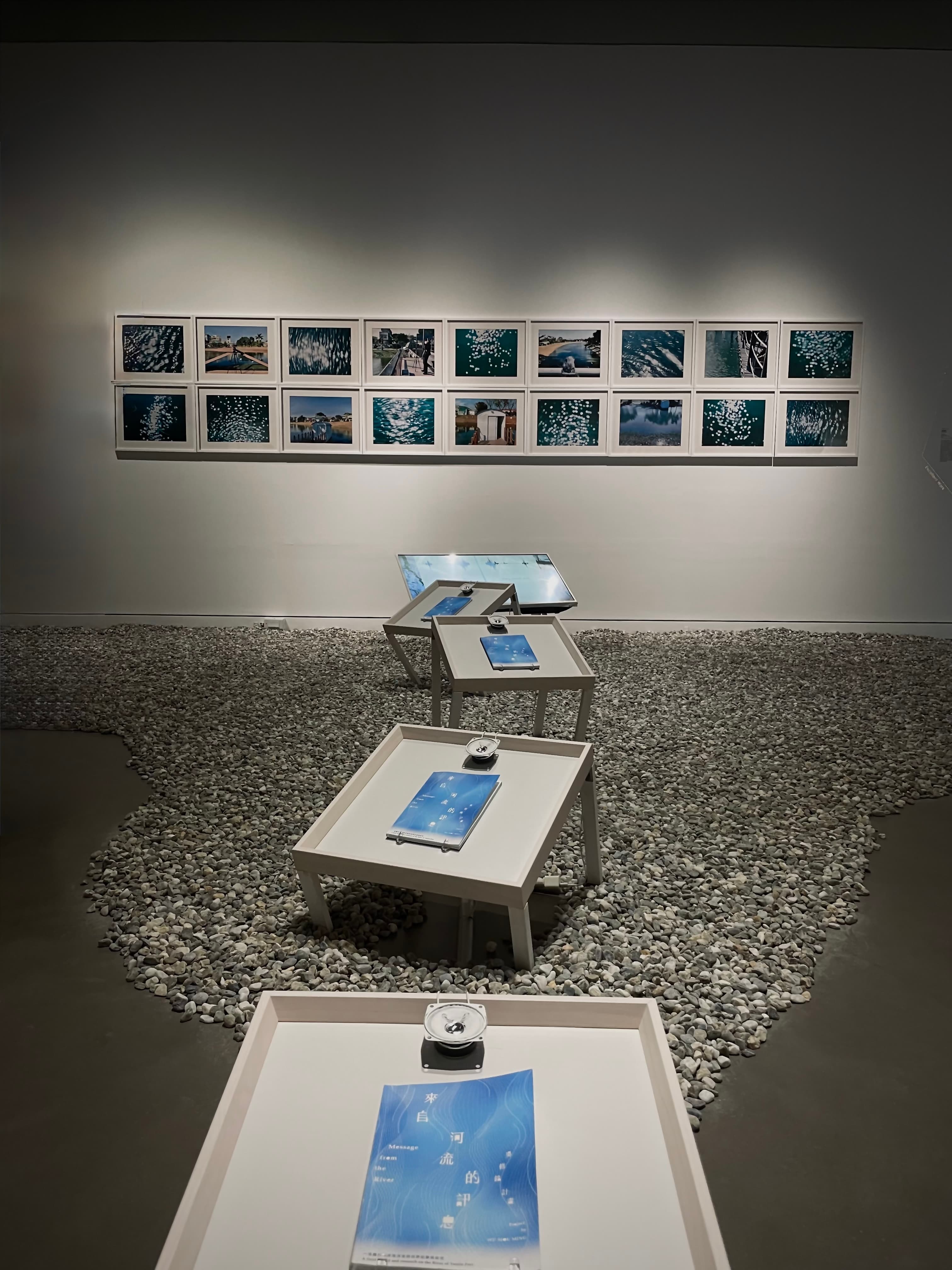高雄市立美術館展覽空間,牆上一排藍色調攝影作品,地面鋪滿白色鵝卵石模擬河床,展桌上放著小音響播放流域錄音