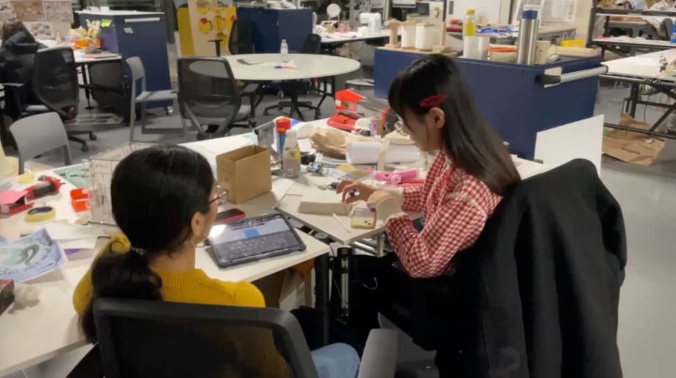 Product Design 工作室裡,兩位組員坐在堆滿材料與工具的桌前組裝原型,一人看著平板